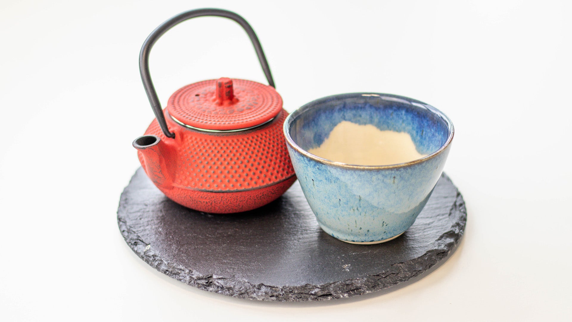 Japanische Eisenteekanne mit einer Keramikteetasse auf einer Schieferplatte. Ocha-Ocha® erklärt Teegeschichte.