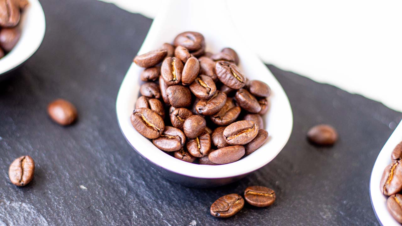 Arabica Kaffeebohnen auf einem Dekolöffel auf einer schwarzen Marmorplatte. Die braunen Bohnen liegen wild verstreut herum.